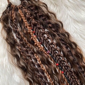 Op de afbeelding: Een bruin haarstuk met een bruine elastiek. Het haar is een mix van golvende en gevlochten strengen in verschillende tinten bruin en beige. Sommige strengen zijn versierd met rode kralen.