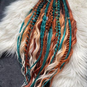 May include: A collection of colorful dreadlocks in shades of teal, orange, brown, and blonde. The dreadlocks are styled with various textures, including braids and wavy sections. The hair extensions are arranged on a white, fluffy surface.