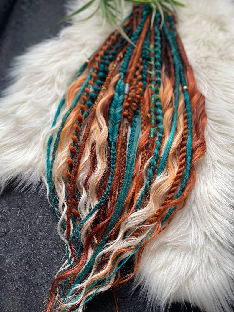 May include: A collection of colorful dreadlocks in shades of teal, orange, brown, and blonde. The dreadlocks are styled with various textures, including braids and wavy sections. The hair extensions are arranged on a white, fluffy surface.