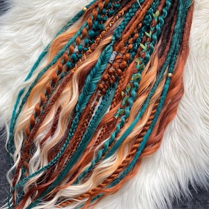 May include: A collection of colorful dreadlocks in shades of teal, orange, brown, and blonde. Some dreads are braided, others are adorned with beads and small green leaves. The dreadlocks are arranged on a fluffy white surface.