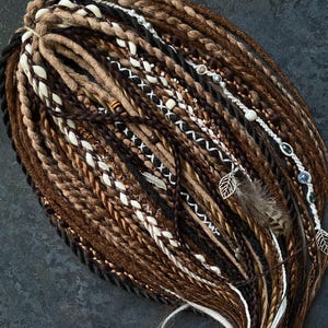 May include: A collection of brown, black, and white dreadlocks, some adorned with decorative beads, feathers, and leaf charms. The dreadlocks are arranged in a layered pattern, showcasing various shades of brown and white.