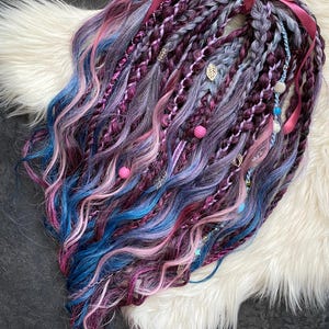 Puede incluir: Una colección de rastas coloridas en tonos morado, azul y rosa, adornadas con cuentas decorativas, abalorios y una cinta rosa. Las rastas están dispuestas sobre una superficie blanca y esponjosa.
