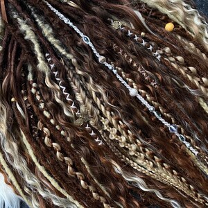 May include: A collection of brown, beige, and blonde dreadlocks, some adorned with decorative beads and charms. The dreadlocks vary in thickness and texture, with some sections braided or wrapped. A few strands are accented with silver and crystal beads.