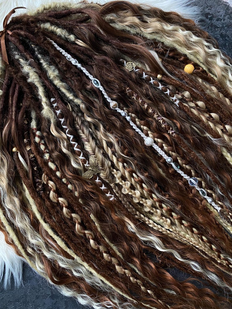 May include: A collection of brown, beige, and blonde dreadlocks, some adorned with decorative beads and charms. The dreadlocks vary in thickness and texture, with some sections braided or wrapped. A few strands are accented with silver and crystal beads.
