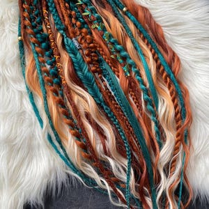 May include: A selection of vibrant dreadlocks in teal, copper, and blonde. The dreads are styled with braids, beads, and small leaf accents, creating a bohemian aesthetic. The hair is arranged on a white, fluffy surface.
