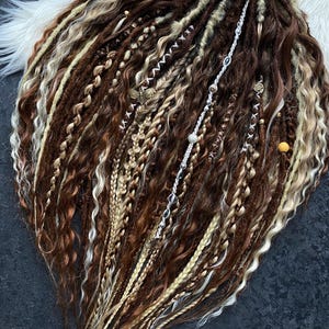May include: A collection of brown, blonde, and light brown dreadlocks, with various textures including braids and wavy sections. Some dreadlocks are adorned with decorative silver and gold accents. The hair is arranged on a white fluffy surface.