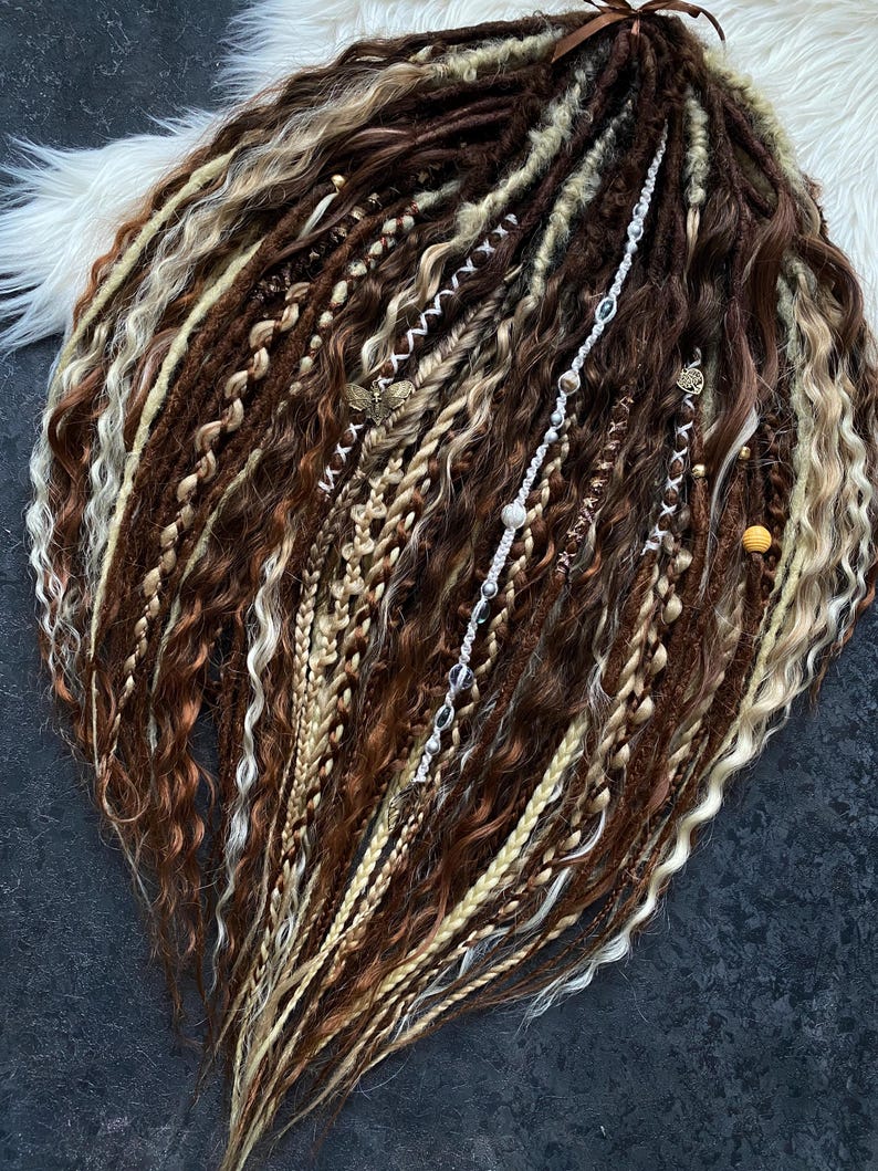 May include: A collection of brown, blonde, and light brown dreadlocks, with various textures including braids and wavy sections. Some dreadlocks are adorned with decorative silver and gold accents. The hair is arranged on a white fluffy surface.