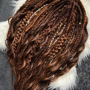 May include: A set of brown and auburn dreadlocks with various braid styles and decorative gold and silver accents. The dreadlocks are arranged on a white faux fur surface, creating a contrast in textures and colors. The hair extensions are styled with intricate details.
