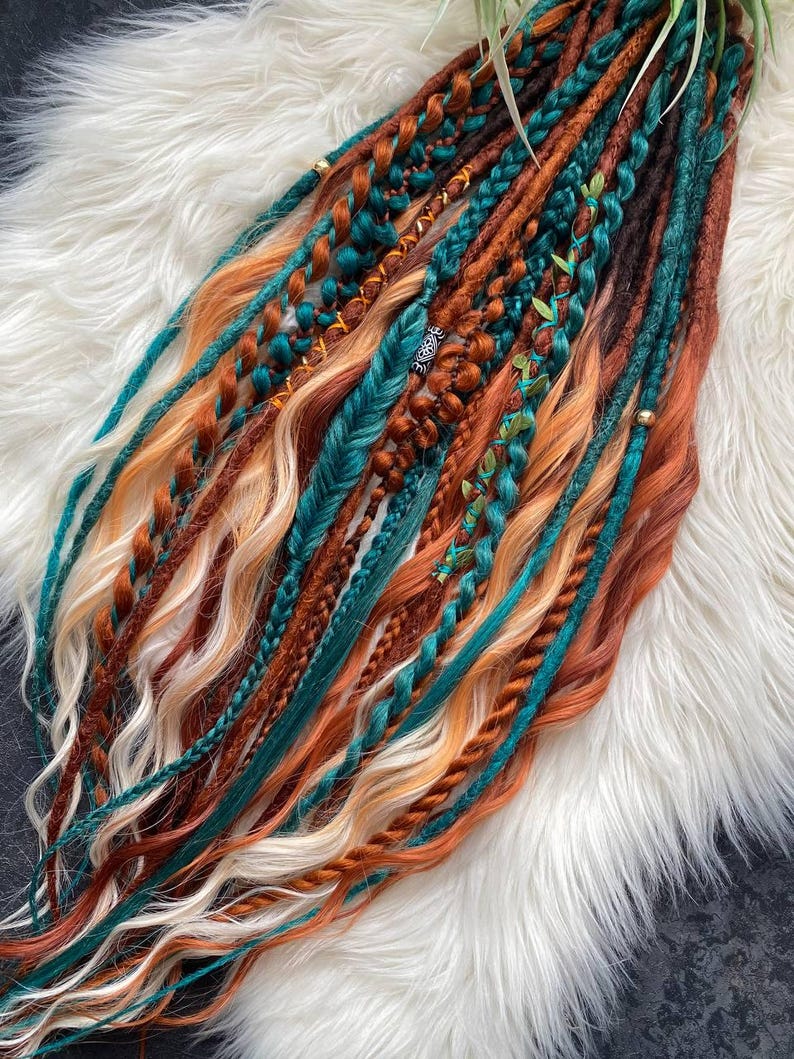 May include: A collection of colorful dreadlocks in shades of teal, orange, brown, and blonde. Some dreads are braided, others are adorned with beads and small green leaves. The dreadlocks are arranged on a fluffy white surface.