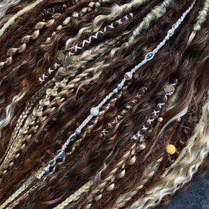 May include: Close-up of brown, blonde, and cream-colored dreadlocks. The dreadlocks are adorned with decorative beads, charms, and wrapped sections. The hair has a mix of textures, including braids and wavy sections, creating a unique and textured look.