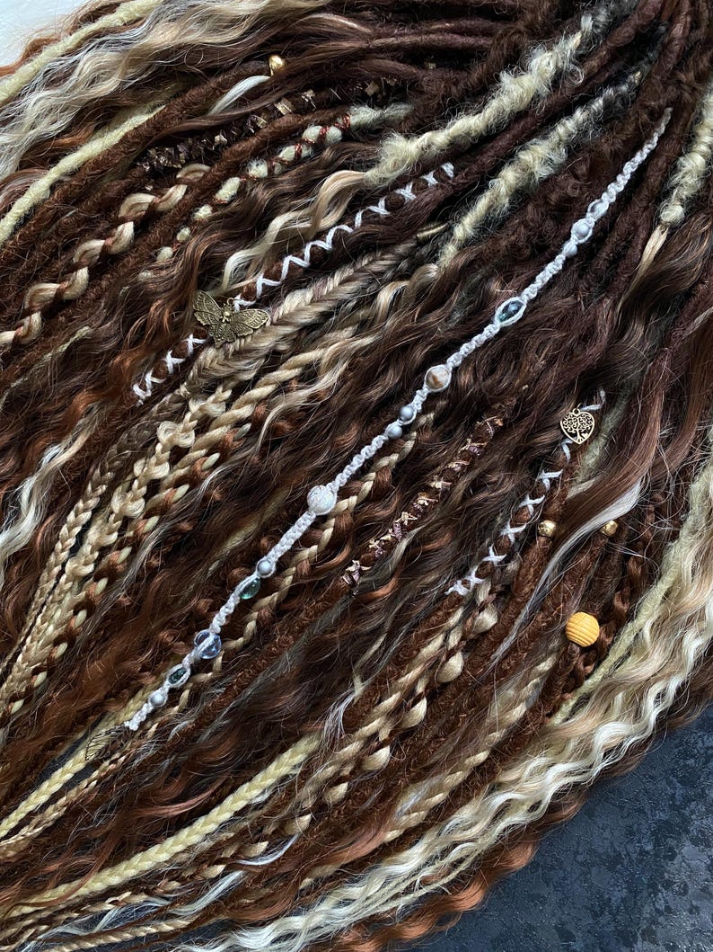 May include: Close-up of brown, blonde, and cream-colored dreadlocks. The dreadlocks are adorned with decorative beads, charms, and wrapped sections. The hair has a mix of textures, including braids and wavy sections, creating a unique and textured look.