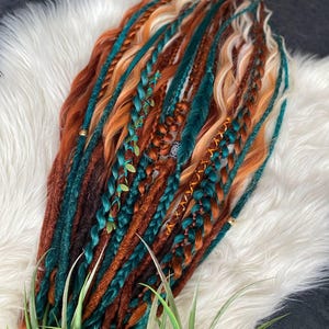 May include: A collection of colorful dreadlocks in shades of brown, orange, teal, and blonde. Some dreads are braided and adorned with small decorative elements. The hair extensions are arranged on a white, fluffy surface.