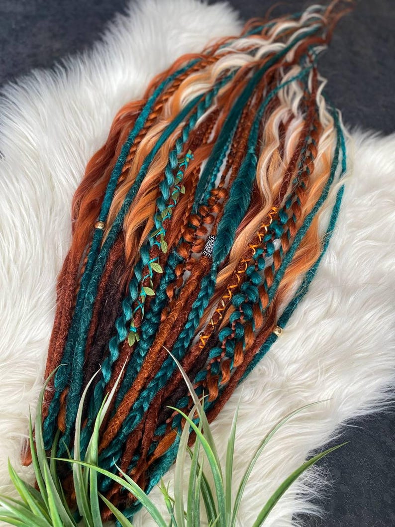May include: A collection of colorful dreadlocks in shades of brown, orange, teal, and blonde. Some dreads are braided and adorned with small decorative elements. The hair extensions are arranged on a white, fluffy surface.