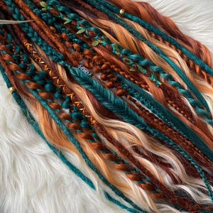 May include: A collection of colorful dreadlocks in shades of teal, brown, orange, and blonde. Some dreads are braided and adorned with small gold beads and green leaf accents. The dreadlocks are arranged on a white, fluffy surface.
