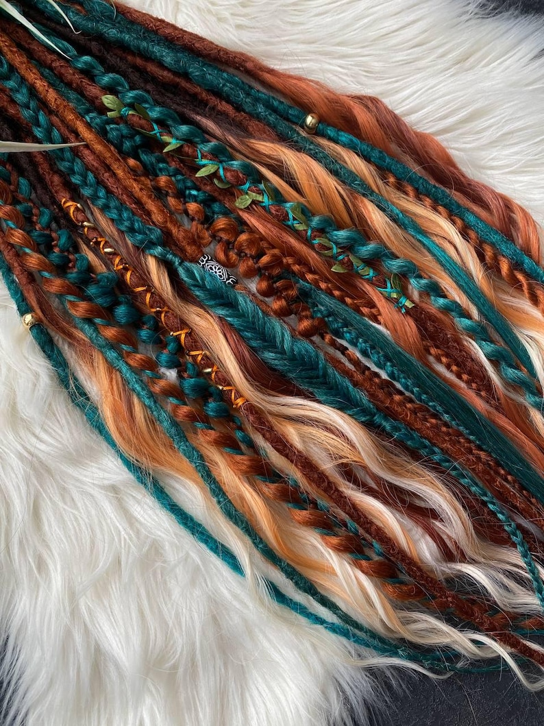 May include: A collection of colorful dreadlocks in shades of teal, brown, orange, and blonde. Some dreads are braided and adorned with small gold beads and green leaf accents. The dreadlocks are arranged on a white, fluffy surface.