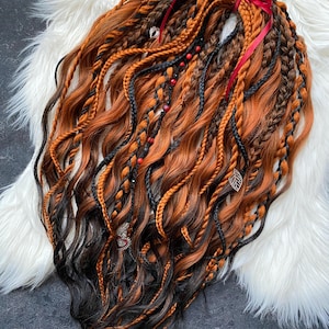May include: A collection of wavy, ombre dreadlocks in shades of brown, orange, and black. The dreadlocks are adorned with small beads, charms, and a red ribbon. The hair is styled on a white, fluffy surface.