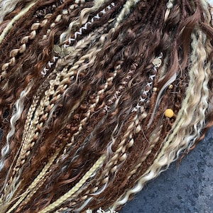 May include: A collection of brown, light brown, and blonde dreadlocks. The dreadlocks are styled with various textures, including braids and wavy sections. Some dreadlocks are adorned with small, decorative charms.