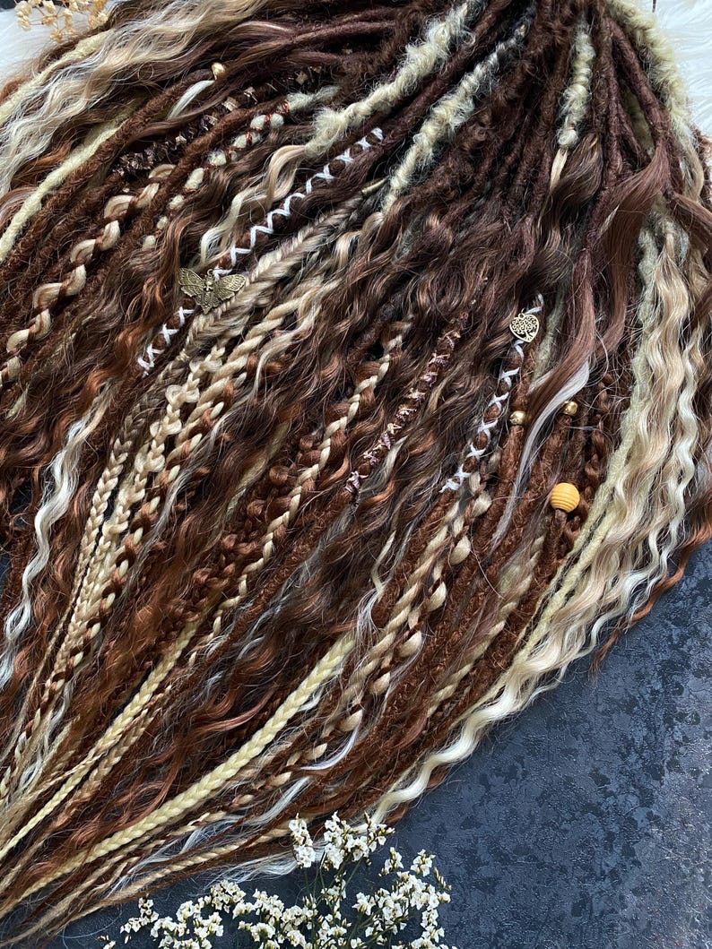 May include: A collection of brown, light brown, and blonde dreadlocks. The dreadlocks are styled with various textures, including braids and wavy sections. Some dreadlocks are adorned with small, decorative charms.