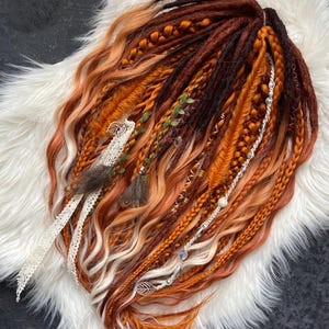 Puede incluir: Conjunto de rastas hechas a mano en tonos naranja, marrón y rubio, con trenzas, plumas y elementos decorativos. Las rastas se presentan sobre una superficie de piel sintética blanca, creando una estética bohemia.