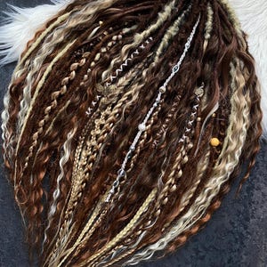 May include: A collection of brown, blonde, and white dreadlocks, some with decorative beads and charms. The dreadlocks are styled with various textures, including braids and wavy sections. A brown ribbon is tied at the top.