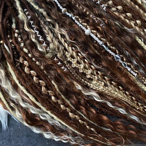 May include: Close-up of brown, blonde, and white dreadlocks with decorative beads and charms. The dreadlocks are styled with various textures, including braids and wavy sections. A butterfly charm is visible.