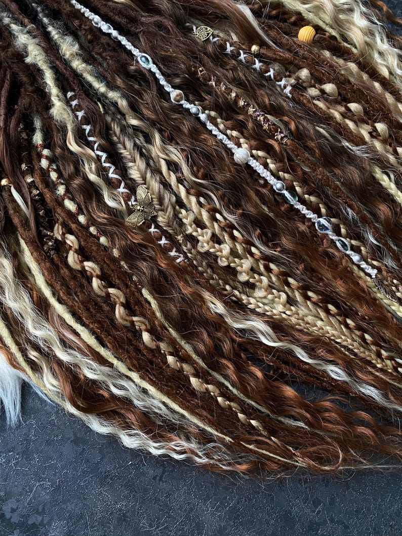 May include: Close-up of brown, blonde, and white dreadlocks with decorative beads and charms. The dreadlocks are styled with various textures, including braids and wavy sections. A butterfly charm is visible.