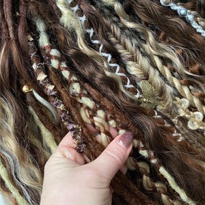 May include: Close-up of brown, blonde, and cream-colored dreadlocks. Some dreads are braided with decorative elements, including a small gold-colored butterfly charm. The dreadlocks have varying textures and shades, creating a natural, textured look.