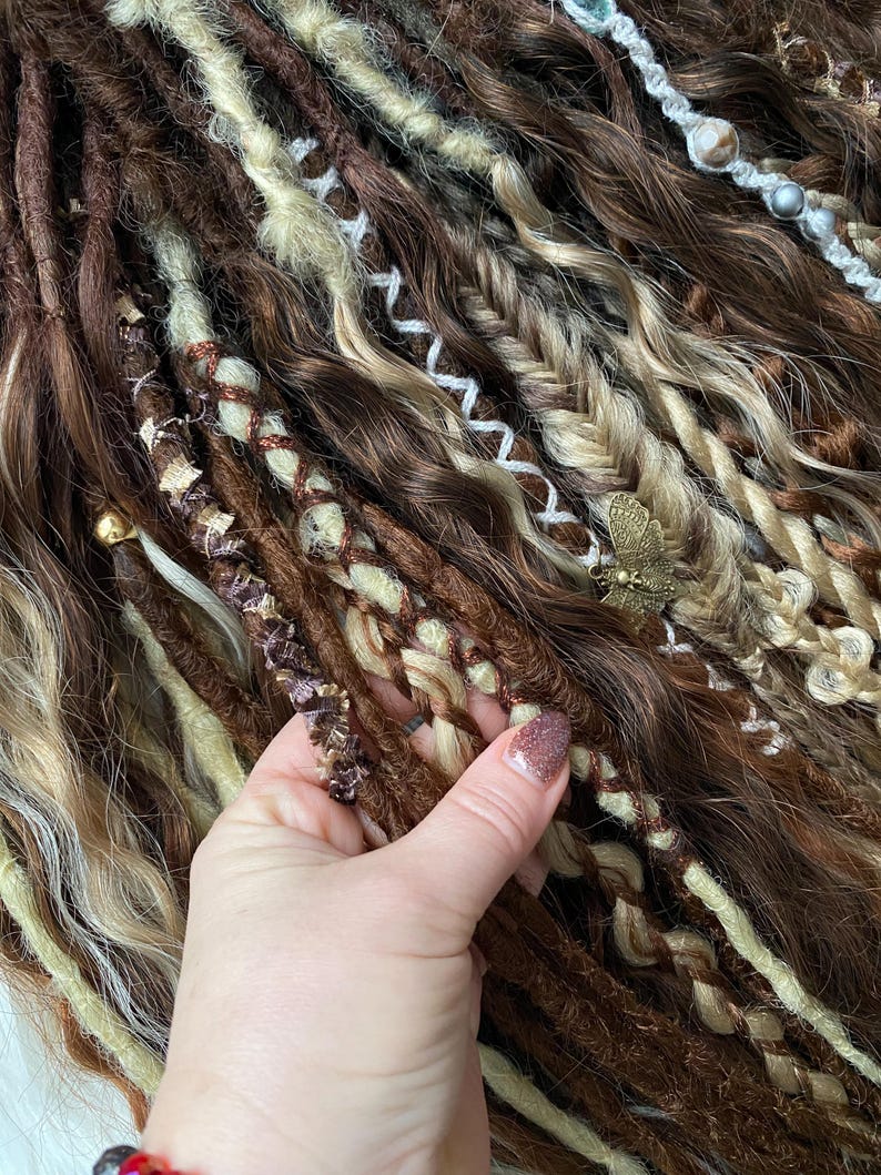 May include: Close-up of brown, blonde, and cream-colored dreadlocks. Some dreads are braided with decorative elements, including a small gold-colored butterfly charm. The dreadlocks have varying textures and shades, creating a natural, textured look.