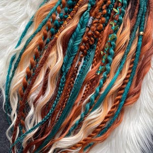May include: A collection of handmade dreadlocks in various shades of brown, teal, orange, and blonde. The dreads are styled with braids, twists, and decorative accents, creating a bohemian aesthetic. The dreadlocks are arranged on a white, fluffy surface.