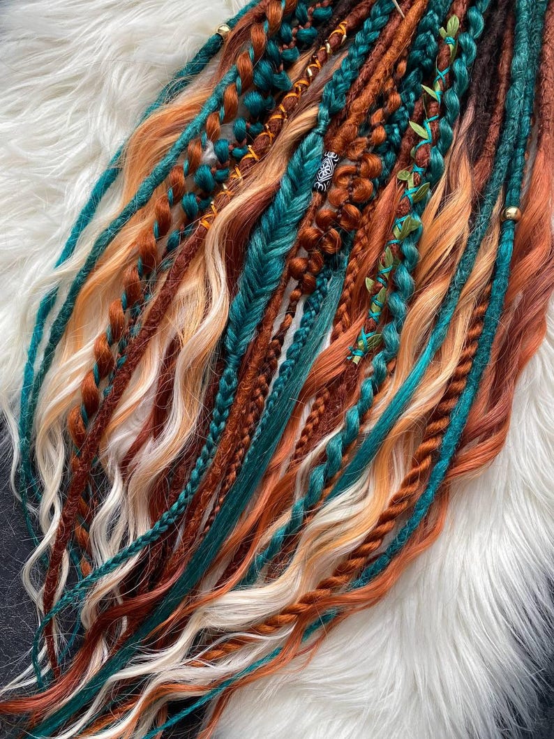 May include: A collection of handmade dreadlocks in various shades of brown, teal, orange, and blonde. The dreads are styled with braids, twists, and decorative accents, creating a bohemian aesthetic. The dreadlocks are arranged on a white, fluffy surface.