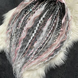 Puede incluir: Un conjunto de rastas sintéticas en tonos grises, blancos y rosas, atadas con una cinta rosa. Las rastas presentan diversas texturas, incluyendo trenzas y secciones onduladas, y están adornadas con cuentas y abalorios. El cabello se muestra sobre una superficie de piel sintética blanca.