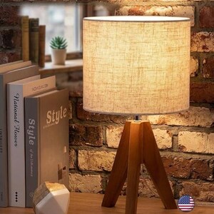 May include: A table lamp with a cylindrical beige fabric shade and a three-legged wooden tripod base. The lamp emits a warm glow, illuminating the surrounding area. The base is constructed from smooth, light-brown wood pieces joined together.