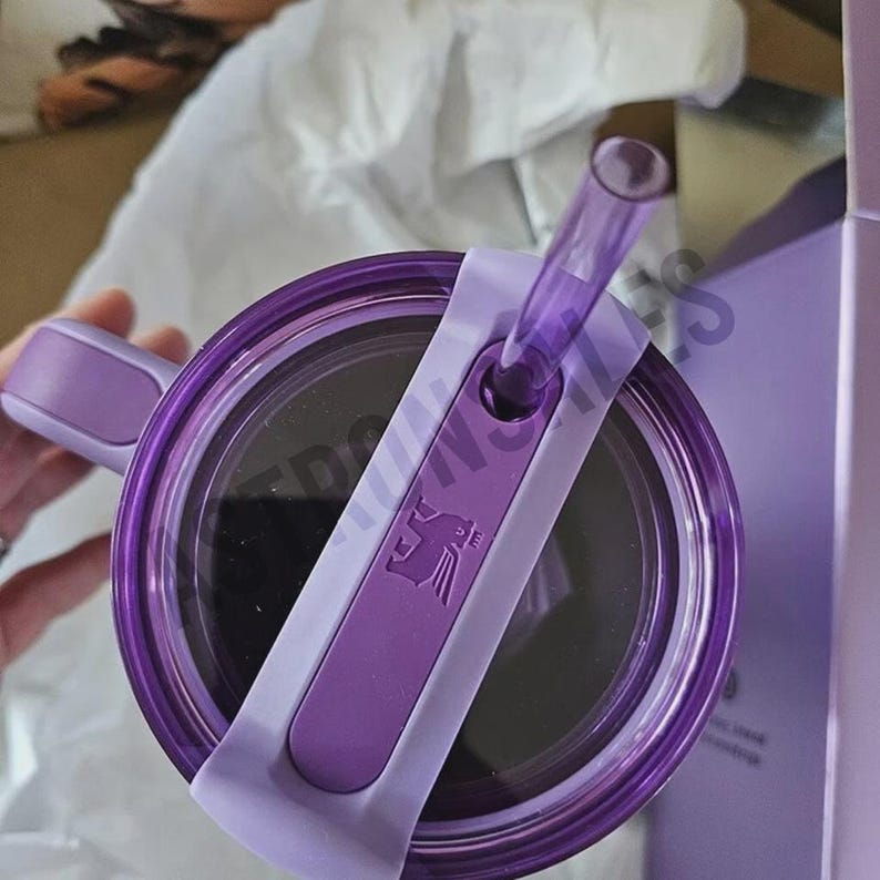 May include: A purple tumbler with a matching lid and straw. The lid features a light purple slider with a logo. The tumbler has a handle and is partially visible, suggesting a travel-friendly design. The overall aesthetic is modern and sleek.