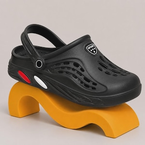 May include: A black clog shoe featuring a perforated upper, a pivoting heel strap, and a side emblem with the text SPORT and three stars. The side of the sole includes a red oval accent and a white oval accent, providing a contrast to the black base.