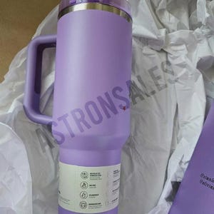 May include: A large, lavender-colored insulated tumbler with a handle. The tumbler has a stainless steel interior and a clear lid. A white label with icons and text is affixed to the side. The tumbler is surrounded by white tissue paper.