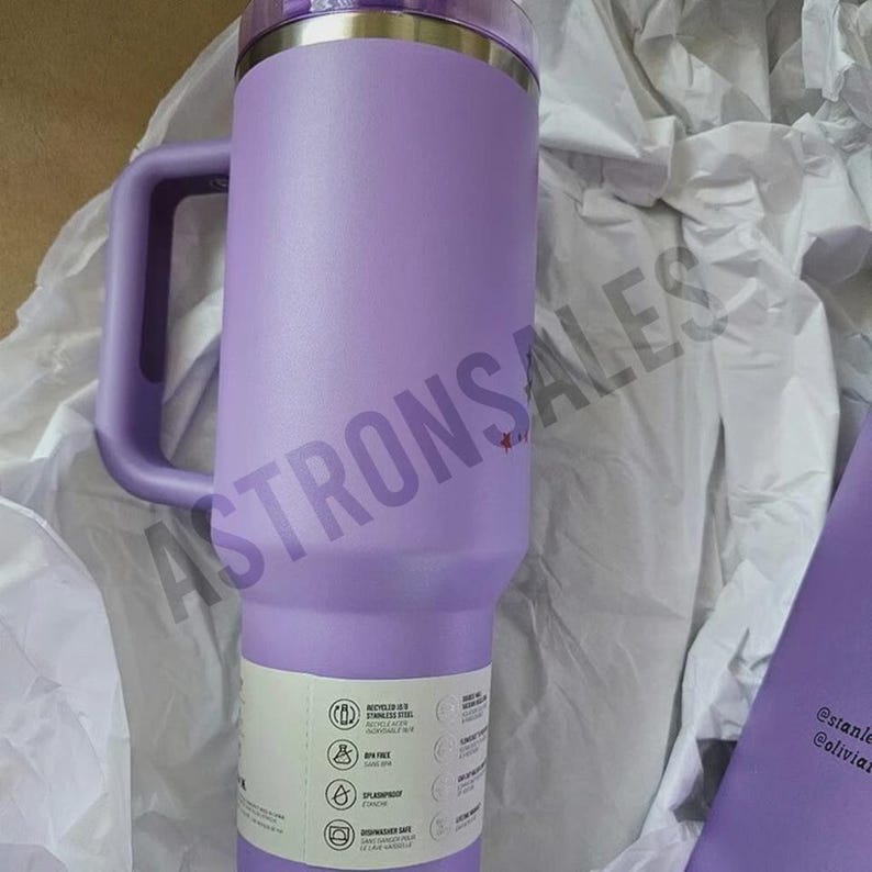 May include: A large, lavender-colored insulated tumbler with a handle. The tumbler has a stainless steel interior and a clear lid. A white label with icons and text is affixed to the side. The tumbler is surrounded by white tissue paper.