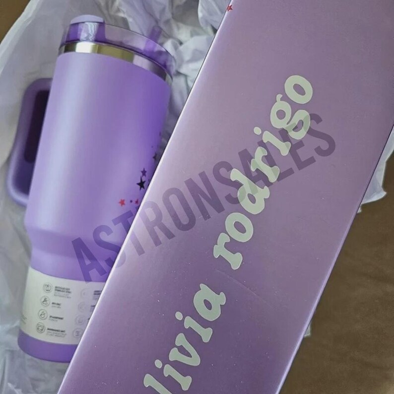 May include: A lavender insulated tumbler with a handle and a lid, next to a matching box. The tumbler has a silver rim and a design of stars. The box has the text "livia rodrigo" and "Astronsales" printed on it.