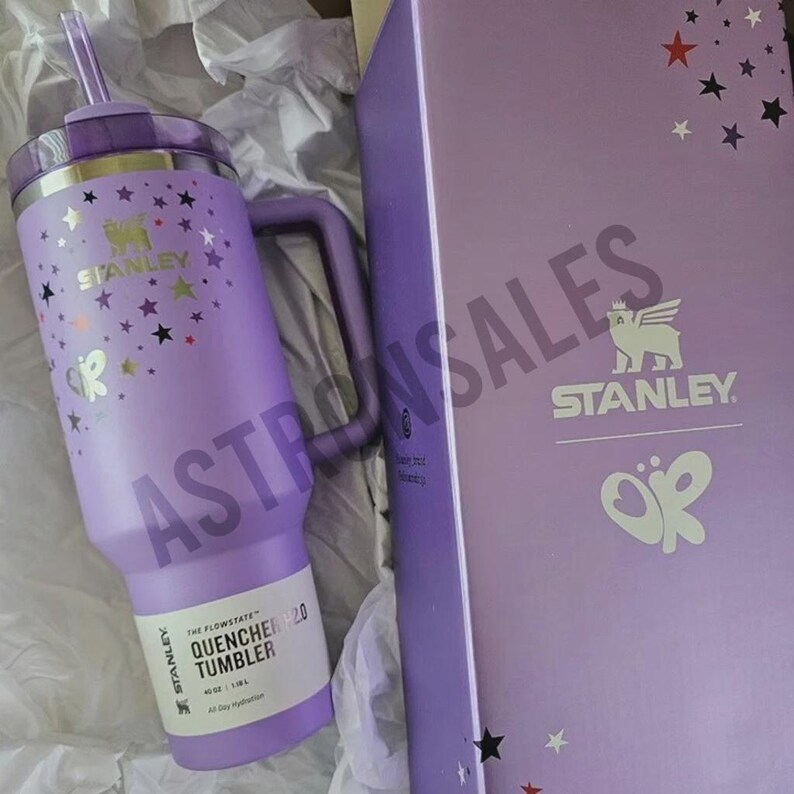 May include: A purple Stanley Quencher 40 oz tumbler with a handle, straw, and lid. The tumbler is decorated with gold stars and the Stanley logo. The box is purple with the Stanley logo and star accents.
