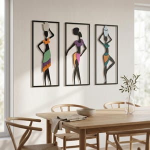 Puede incluir: Conjunto de tres obras de arte mural que representan siluetas negras de mujeres que llevan vasijas sobre sus cabezas. Cada figura está enmarcada en un borde rectangular negro y adornada con ropa colorida en tonos naranja, morado, verde y azul.