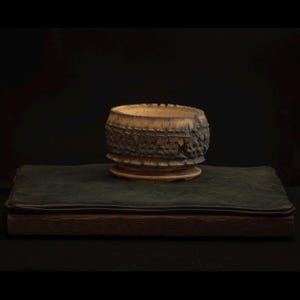 Può includere: Una ciotola in ceramica fatta a mano con un esterno marrone scuro strutturato e un interno marrone più chiaro. La ciotola poggia su una piccola base rotonda ed è esposta su una superficie verde scuro su sfondo nero. Il design della ciotola suggerisce un'estetica rustica o artigianale.