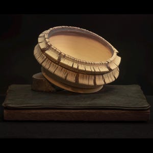 Może przedstawiać: Ręcznie wykonana, jasna ceramiczna miska z unikalnym, teksturowanym wzorem obręczy. Misa jest podparta małą, okrągłą drewnianą podstawą i spoczywa na ciemnozielonej, skórzanej książce. Tło jest czarne.