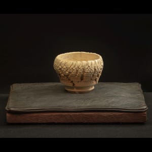 Può includere: Una piccola ciotola in ceramica strutturata, bianco sporco, con un'ampia apertura, posta su una base in legno rivestita in pelle grigio scuro. L'esterno della ciotola presenta un motivo ruvido e organico. La base ha un bordo in legno marrone scuro.