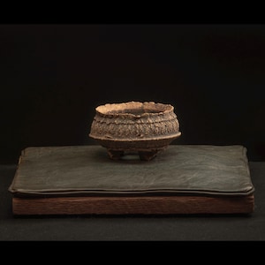 Peut inclure: Un petit bol en céramique marron texturé avec trois petits pieds, posé sur un livre relié en cuir vert foncé. Le bol a une texture rugueuse et organique et un large dessus ouvert. Le livre a une base en bois.