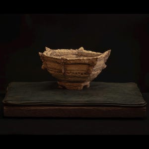 Può includere: Una ciotola in ceramica beige strutturata con un bordo irregolare, appoggiata su una superficie verde scuro. La ciotola ha un aspetto ruvido e organico, con creste visibili e una forma leggermente angolare. Lo sfondo è nero.