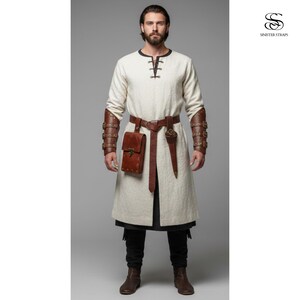 May include: A cream-colored tunic with long sleeves, a black neckline, and brown leather accents. The outfit includes a brown leather belt, a pouch, and arm guards. A dagger is attached to the belt. The outfit is completed with black pants and brown boots.