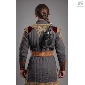May include: A grey quilted tunic featuring gold embroidered patterns on the shoulders and hem, paired with a brown leather belt and brown leather bracers. A black leather scabbard with two buckles holds a silver-toned dagger on the back.