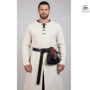 May include: A cream-colored tunic with long sleeves and a black leather belt. A black leather pouch with a brown leaf design is attached to the belt. The tunic has a black lace-up neckline. The brand logo "SINISTER STRAPS" is in the upper right corner.