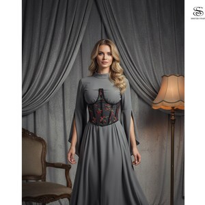 May include: A gray dress with long sleeves and a high neckline. The dress features a black corset with red and green floral embroidery. The model is standing in front of a gray curtain and a vintage chair.
