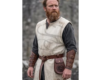 Peut inclure: Une tunique sans manches blanc cassé portée sur une chemise à manches longues gris foncé, associée à une ceinture en cuir marron avec une petite pochette et des brassards en cuir marron assortis dotés de trois boucles en laiton.