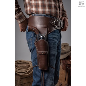 May include: A wide brown leather gun belt featuring a large silver-toned metal buckle and an integrated holster containing a revolver with a dark wooden grip. The belt is worn over blue denim jeans and a brown and white plaid long-sleeve shirt.